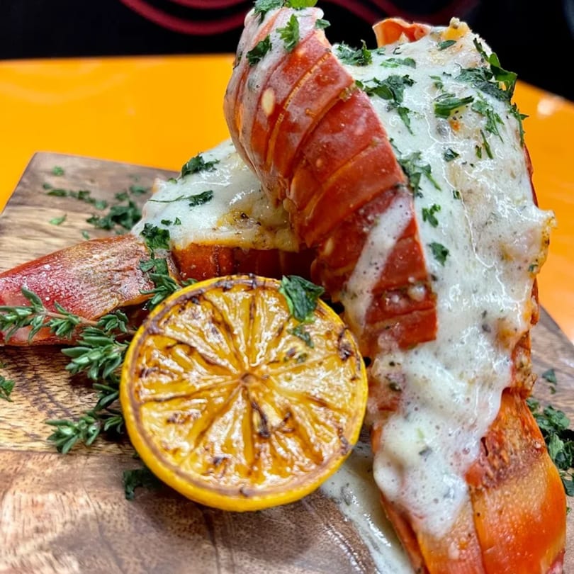 Two lobster tails with white sauce and herbs, served with a grilled lemon on a wooden board.