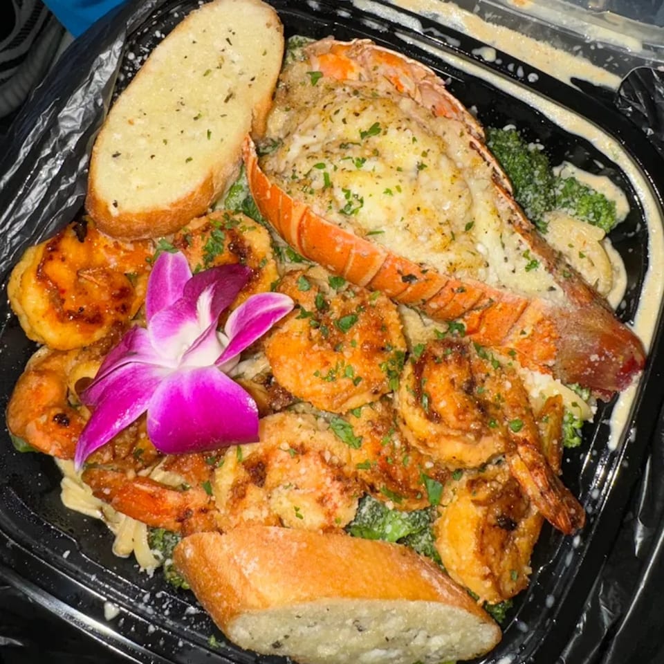 A Caribbean seafood dish with lobster tail, shrimp, pasta, broccoli, garlic bread, and a purple orchid.